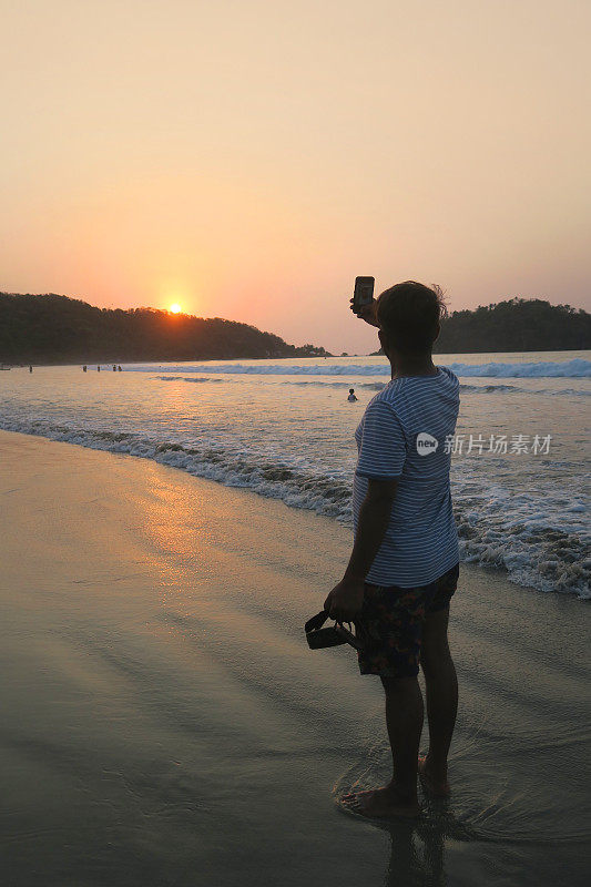 20岁出头的亚洲印度游客在海滩上自拍，手持手机，自拍男子在度假果阿海滩用手机自拍日落，t恤，沙子，海边日落/日出的剪影